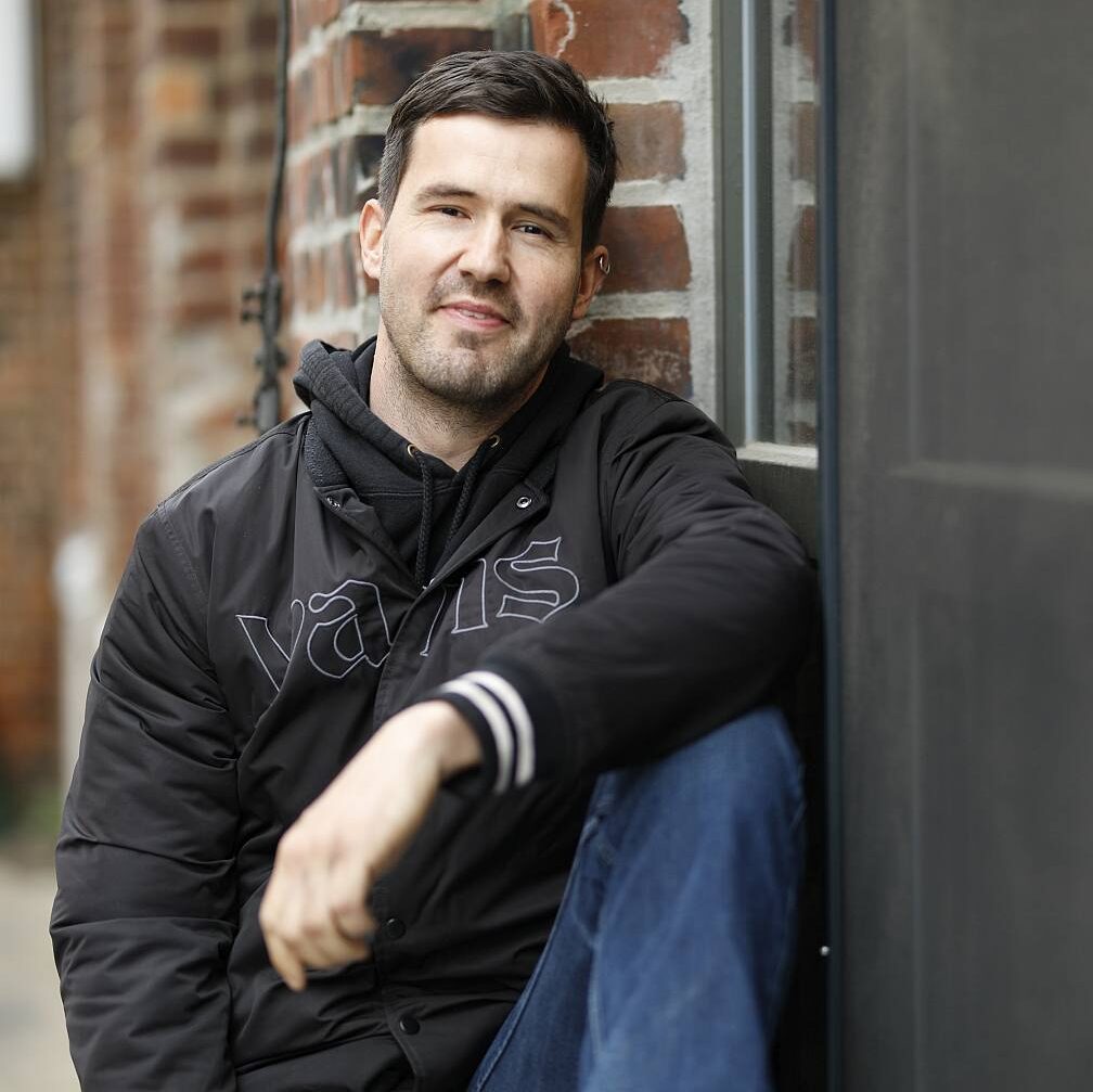 Autor Constantin Dupien sitzt entspannt auf einer Fensterbank und lächelt in die Kamera. Er hat einen Fuß auf dem Sims abgestellt.